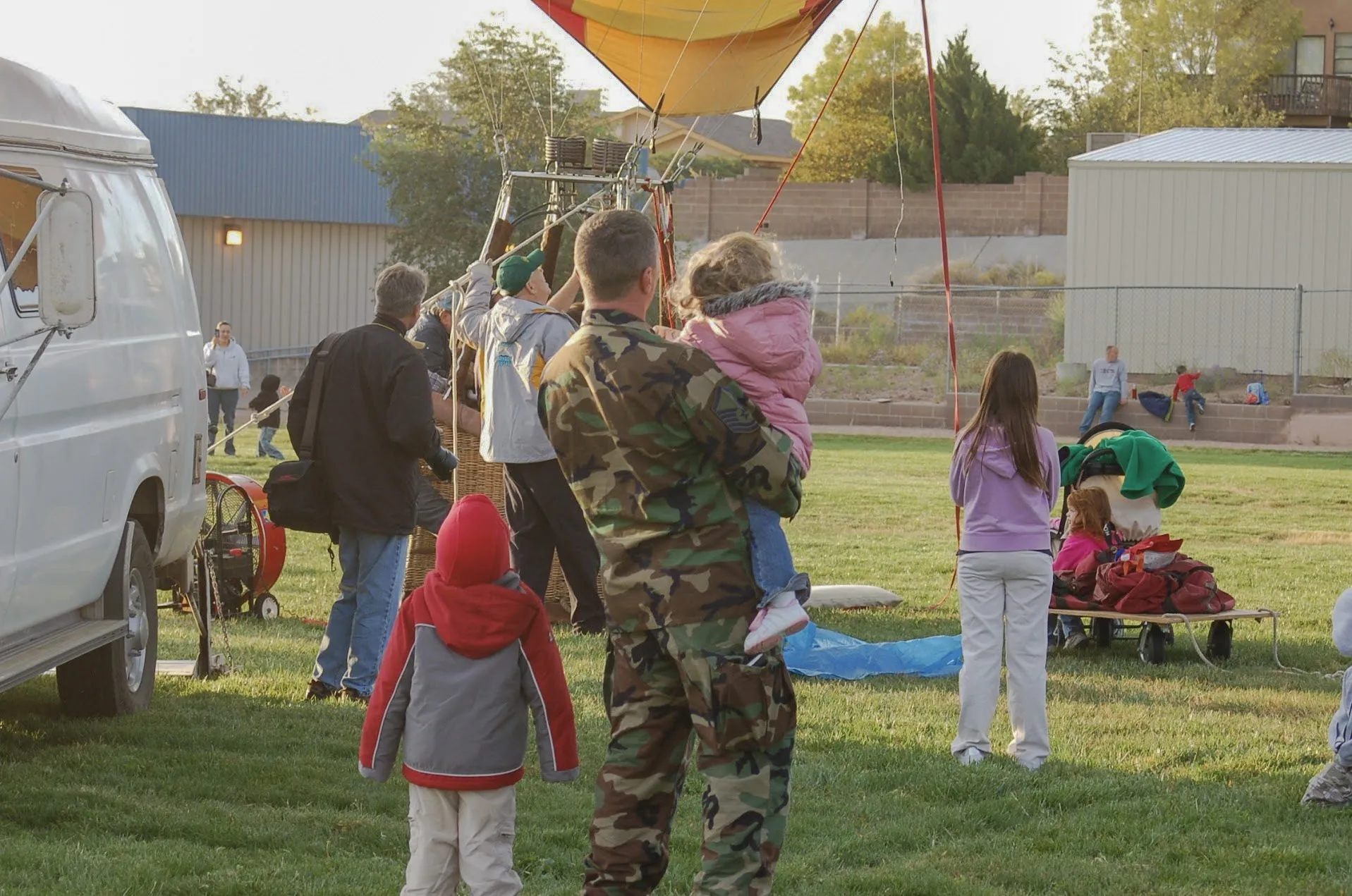 Corey Frasure in uniform holding a child on his arm, watching a hot-air balloon inflate at a school field
