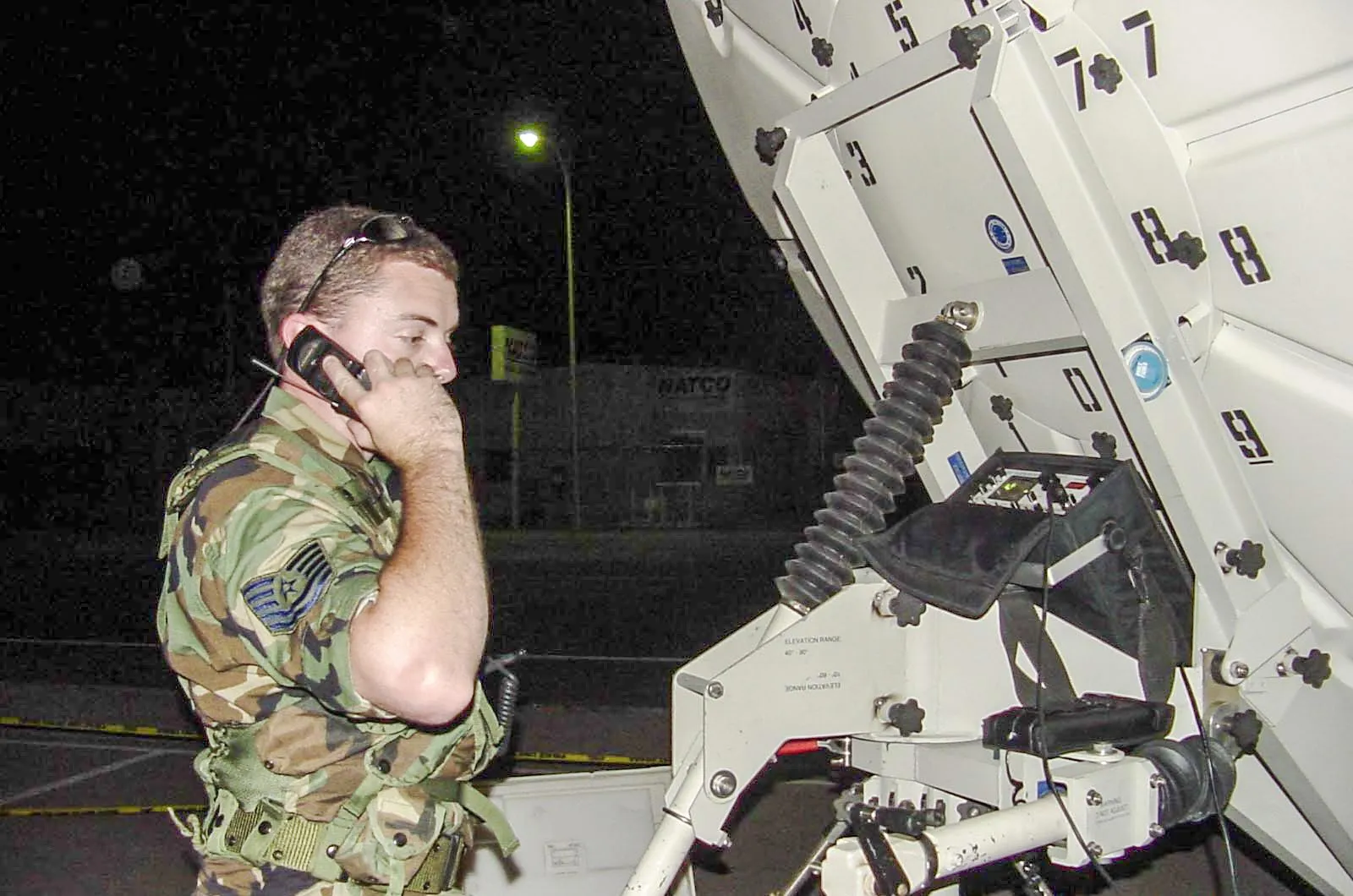 Corey Frasure in uniform at night, on a handset beside a satellite terminal